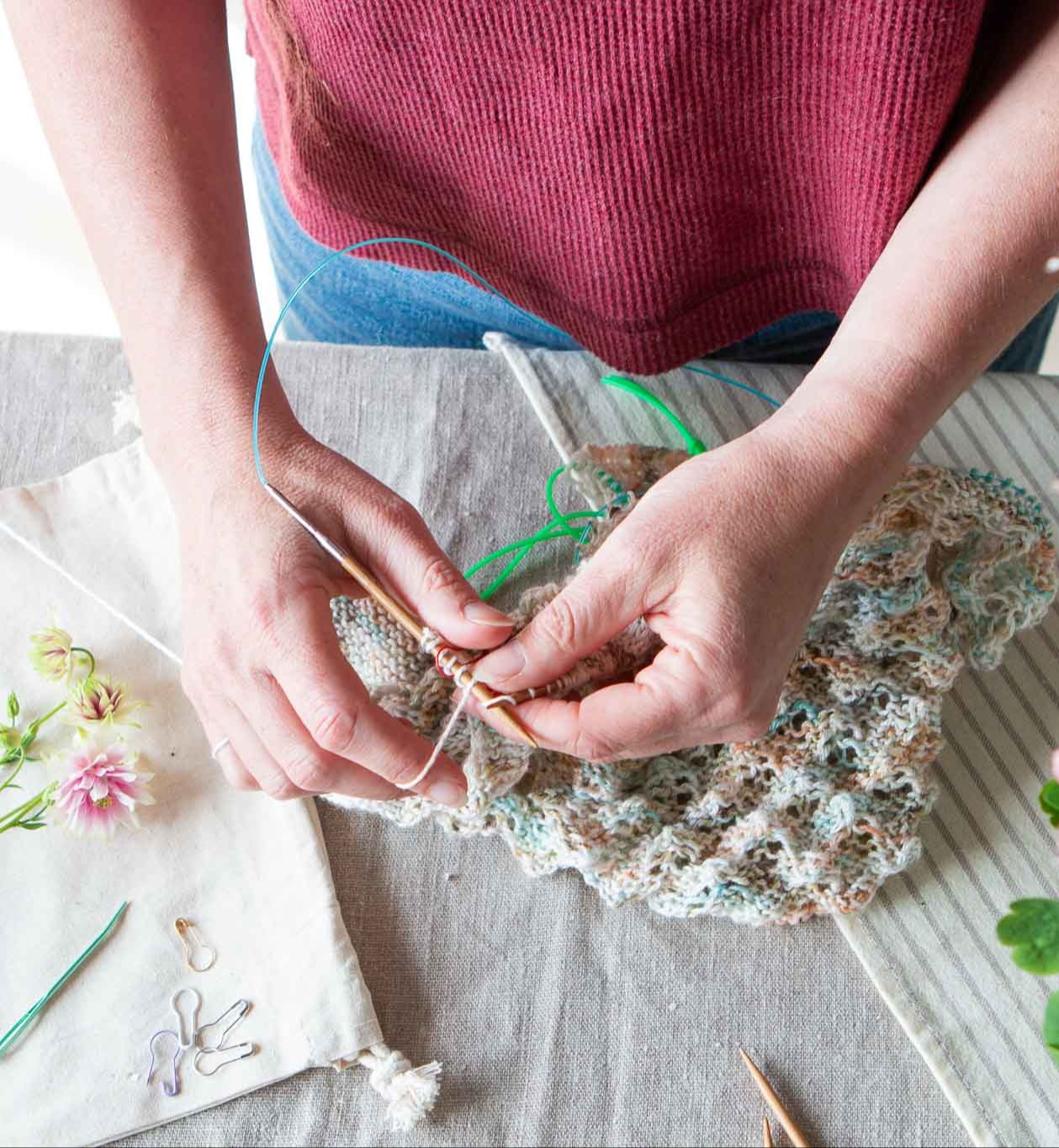 Two hands holding a set of wooden knitting needles with a lace knitting project on them sorrounded by summer flowers.