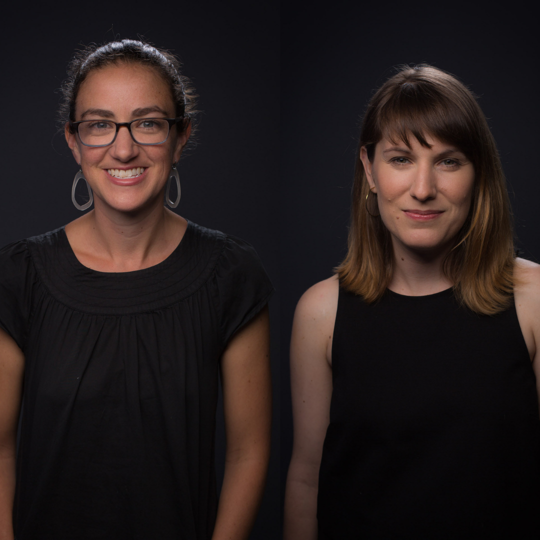 An image of two white women. The woman on the left has brown hair that is tied back, and is wearing brown glasses, large hoop earrings, and a black t-shirt. Woman on the right has light brown hair, short bangs and shoulder-length hair, and is wearing a black tank top.