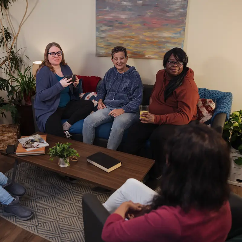 A group of five people gather in a living room while actively listening to one of them.