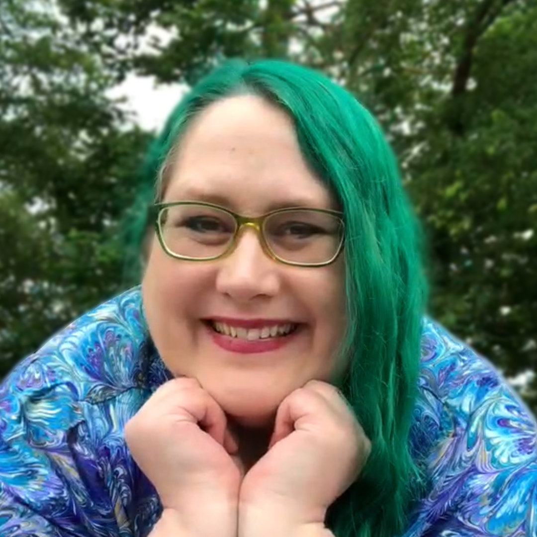 author Veronica Bartles, smiling in front of a background of trees, with green hair and wearing a shirt with swirls of blue, purple, green, and yellow like peacock feathers