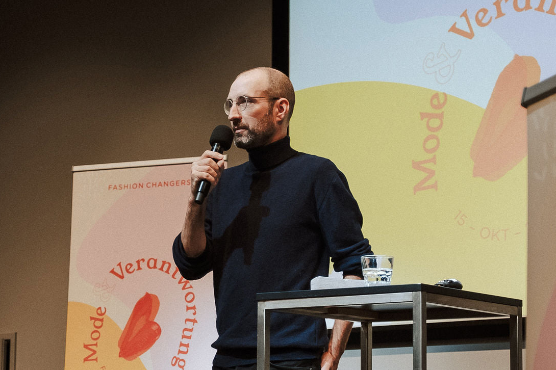 Mann mit Rollkragen Pullover und Brille steht auf der Fashion Changers Konferenzbühne und hält einen Vortrag