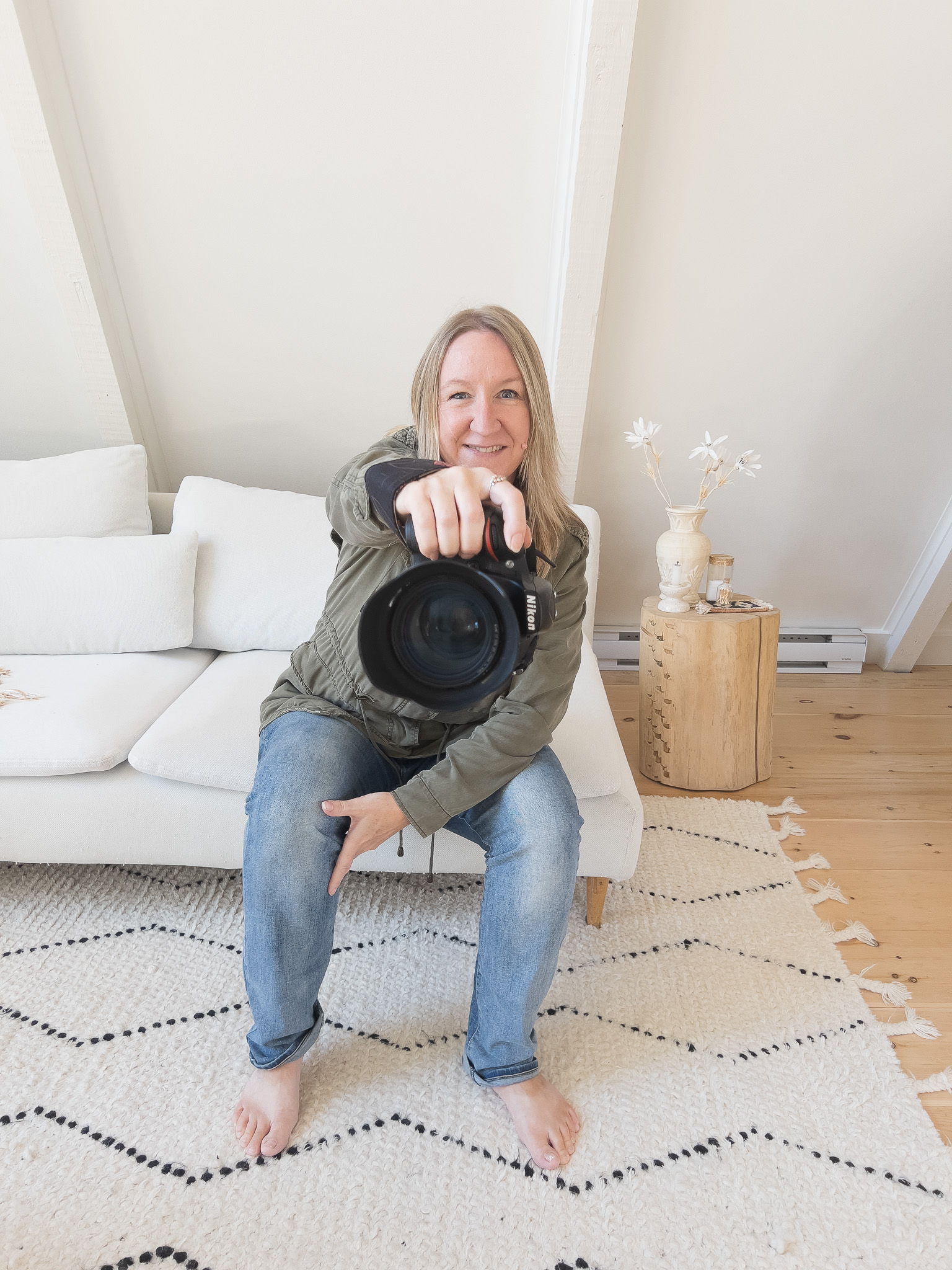 female photographer with blond hair, wearing olive green jacket and blue jeans, sitting on white sofa, holding out camera with large lens