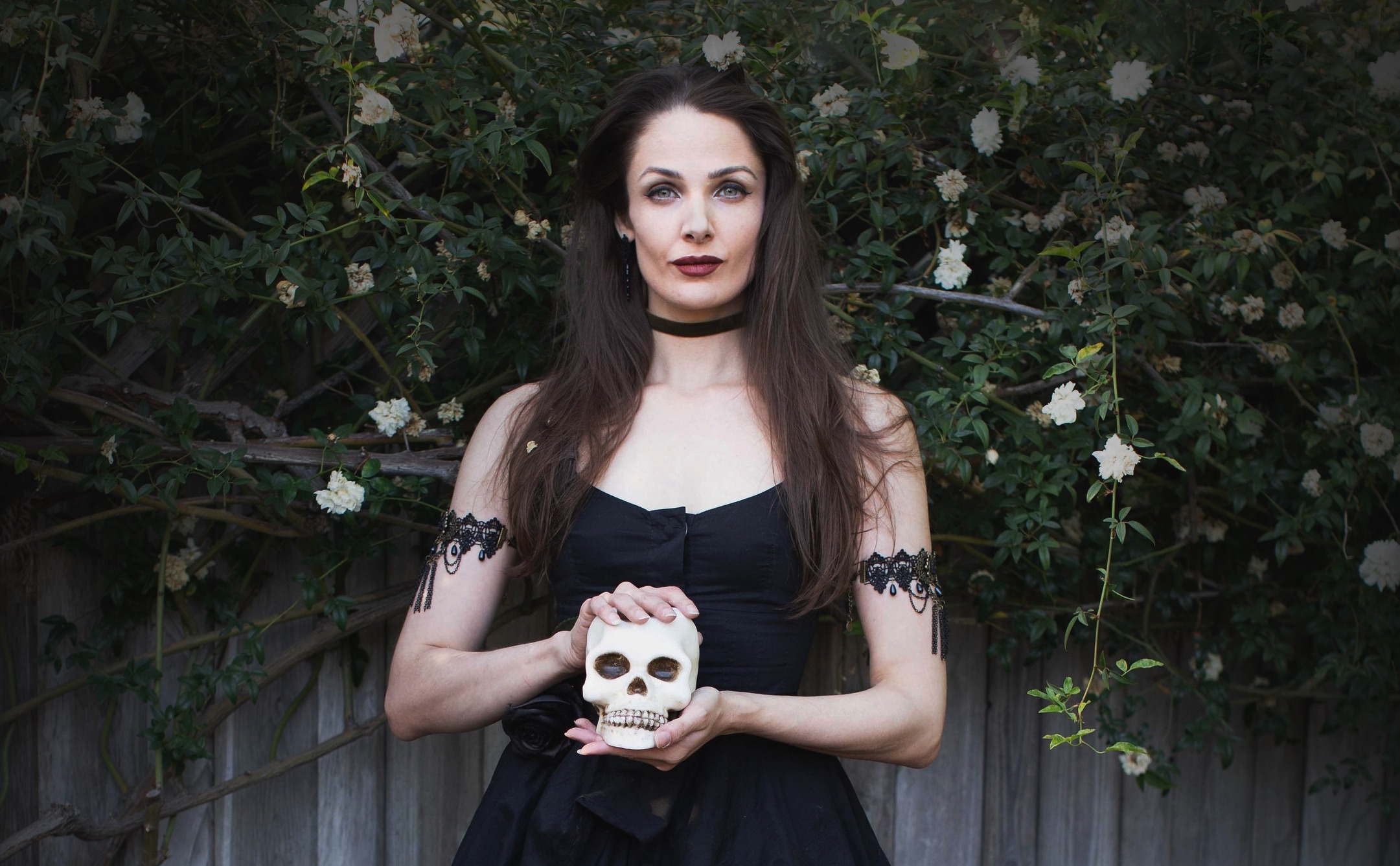 Arden Leigh wearing a black dress with a black choker and armbands, holding a skull. They are standing in front of a wall of roses.