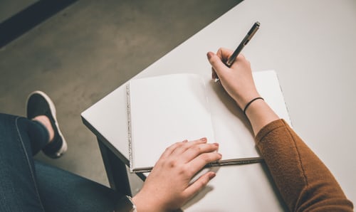 An open notebook on a desk with a hand holding a pen over it