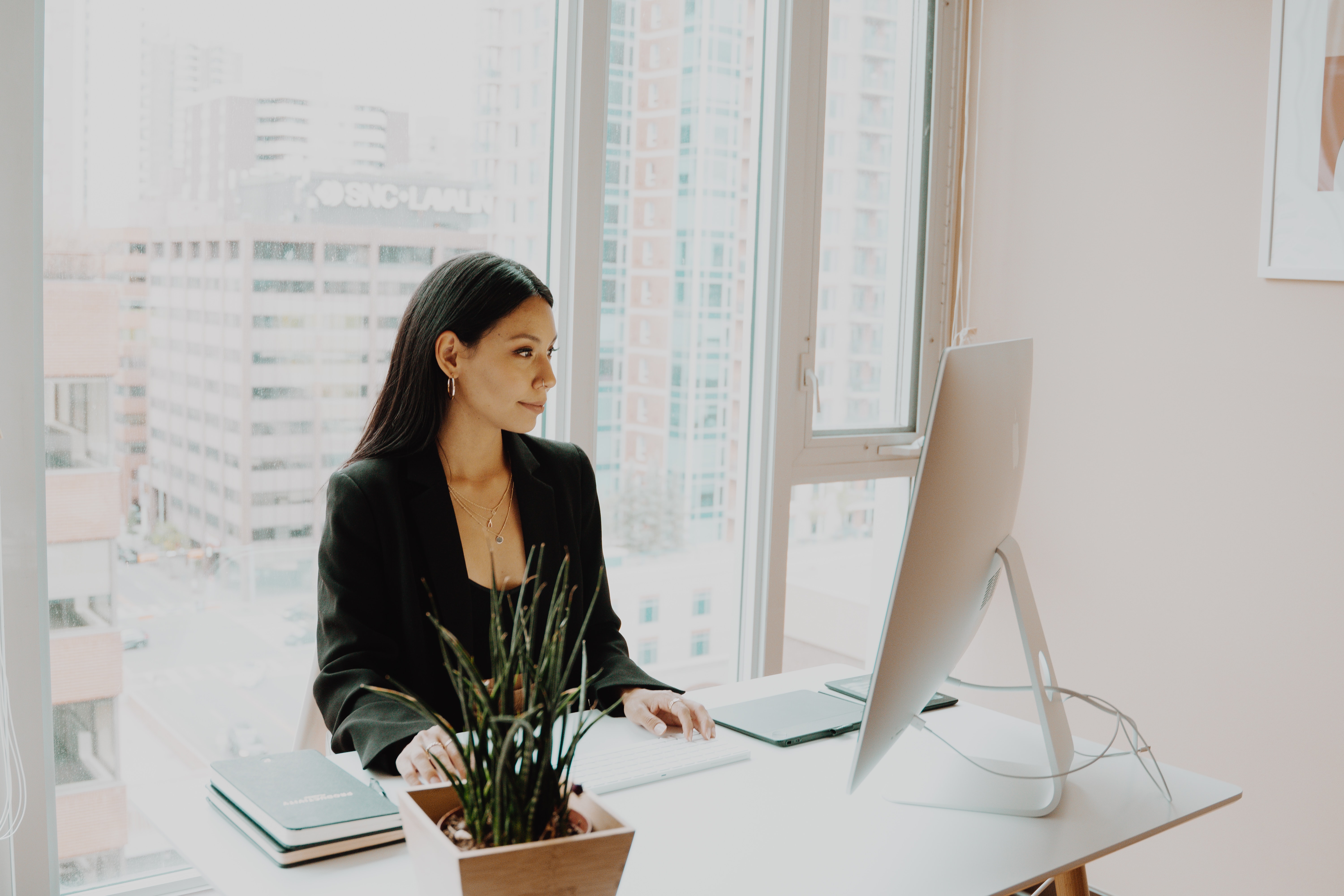 woman sits at a desk with computer with window and city scape through windows behind her