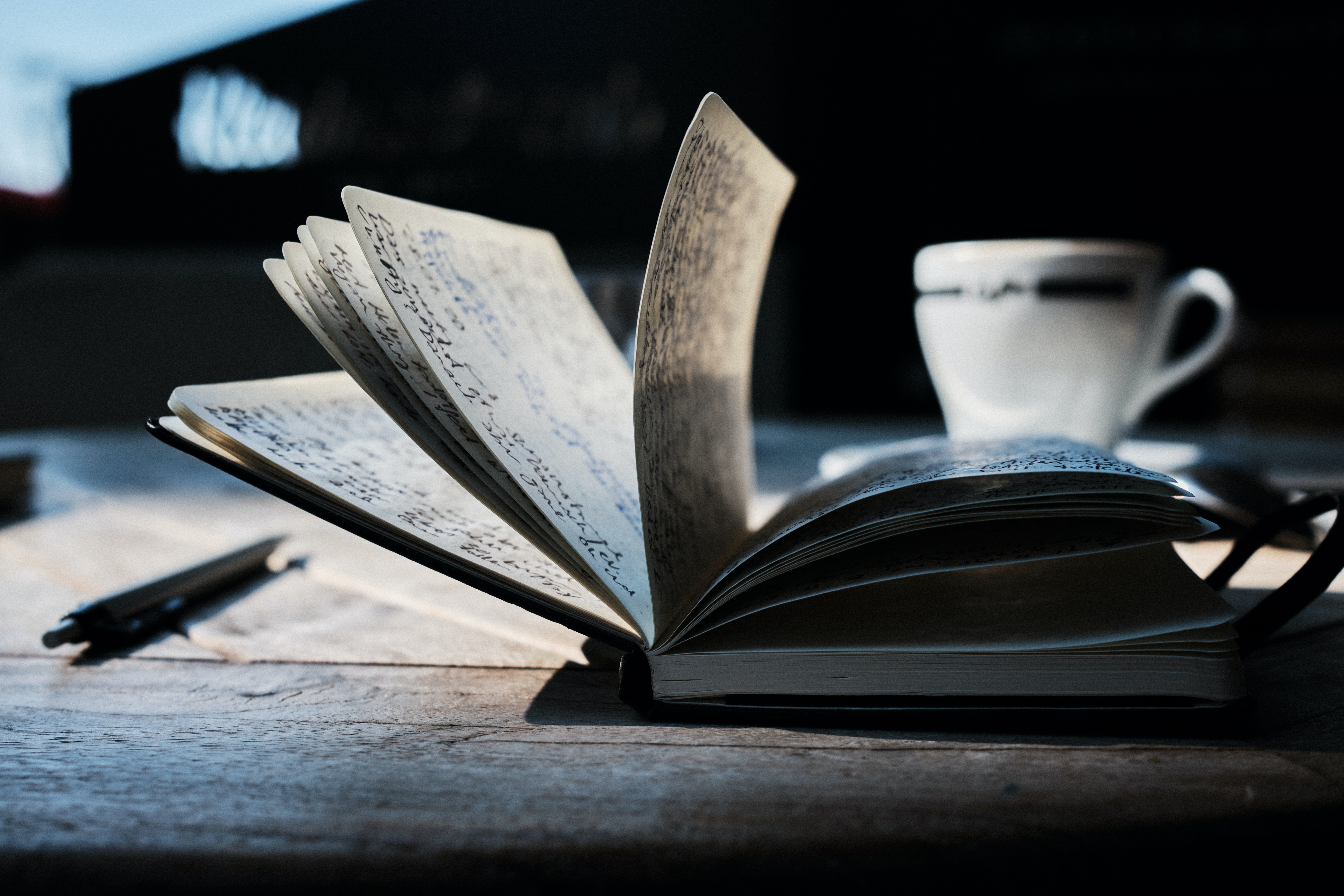 Journal open with writing visible next to a black pen with a white coffee cup in the back ground