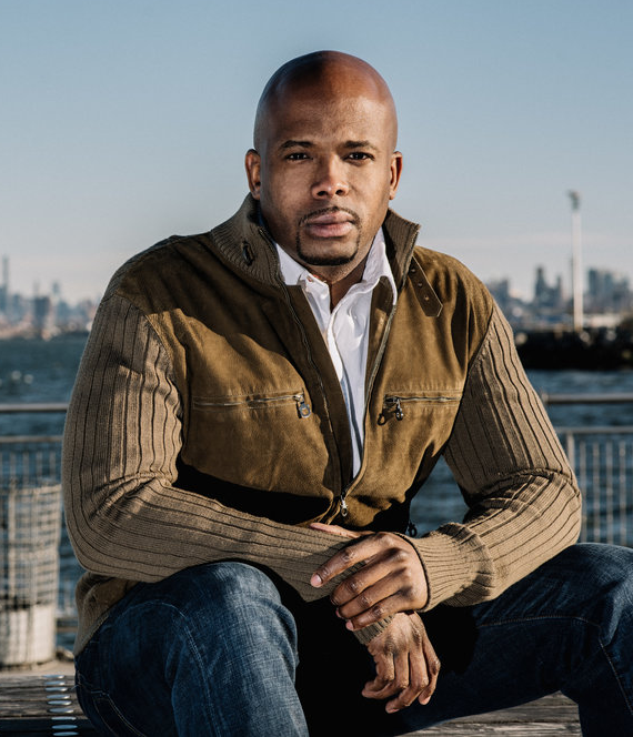 Picture of Chesney Snow, a middle aged Black man, sitting and looking at the camera with a determined expression. He is wearing a brown suede and fabric jacket, a white button down shirt, and jeans. His head is shaved, he has facial hair, and he sits with his elbows resting on his knees in front of a river. There is a city skyline behind him.