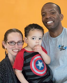 Kate Feinberg Robins and DeShawn Jamar Grim Robins smile and hold our two-year-old son in front of an orange wall at our studio in 2018