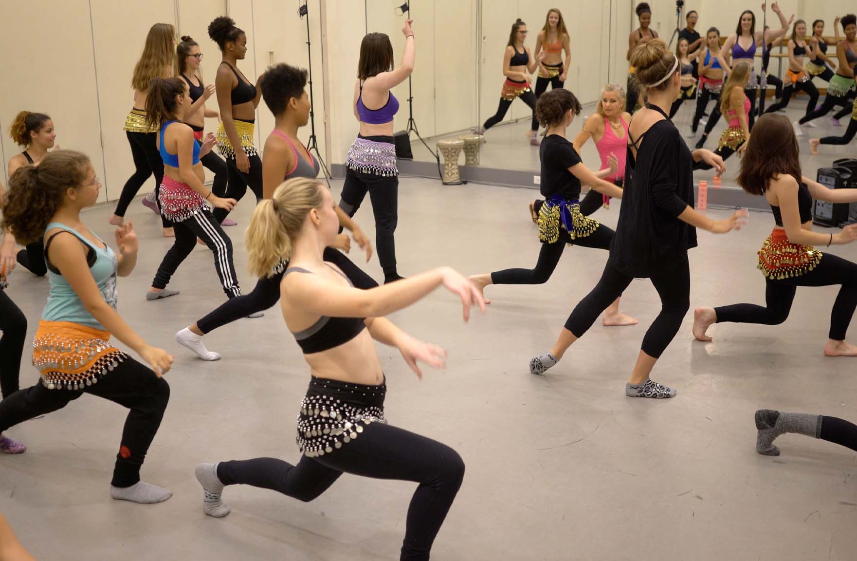 young and mature women wearing hip scarfs learning to belly dance in studio