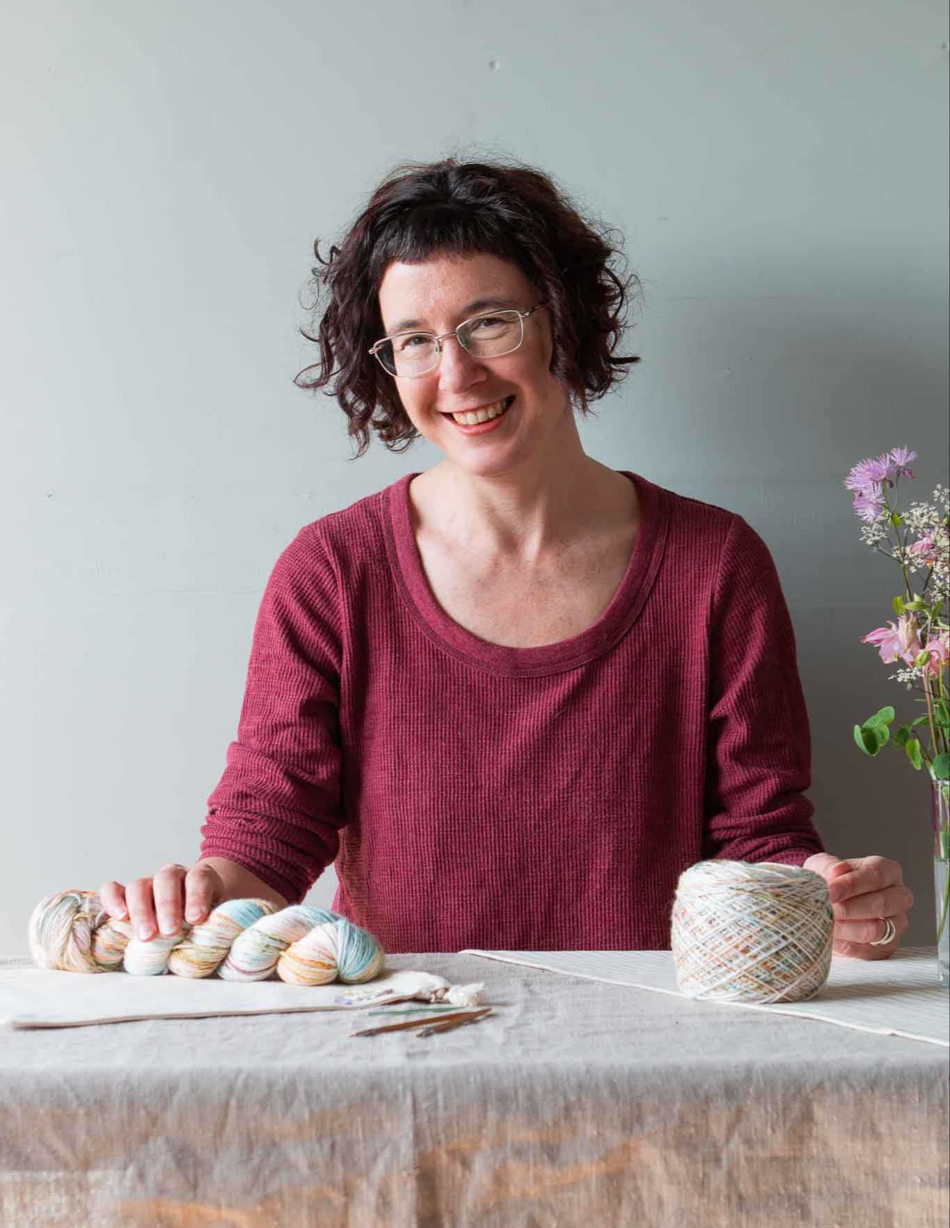 Woman sat behind a table smiling at the camera with a skein of hand-dyed yarn in her right hand.