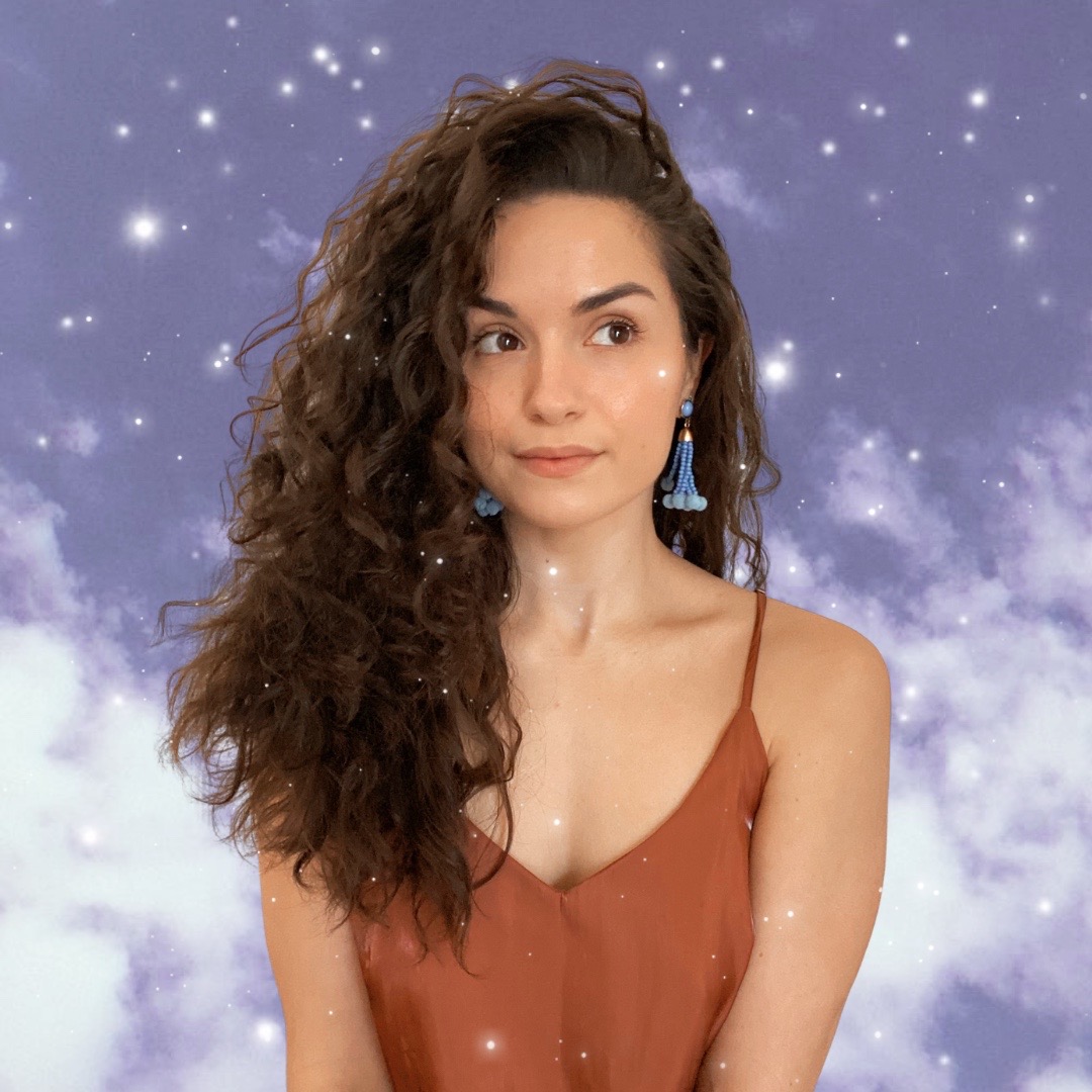 Maria is wearing her hair down, with purple earrings and the tops of her shoulders are seen. Her eyes are looking to the right and there is a dreamy cloud background. 