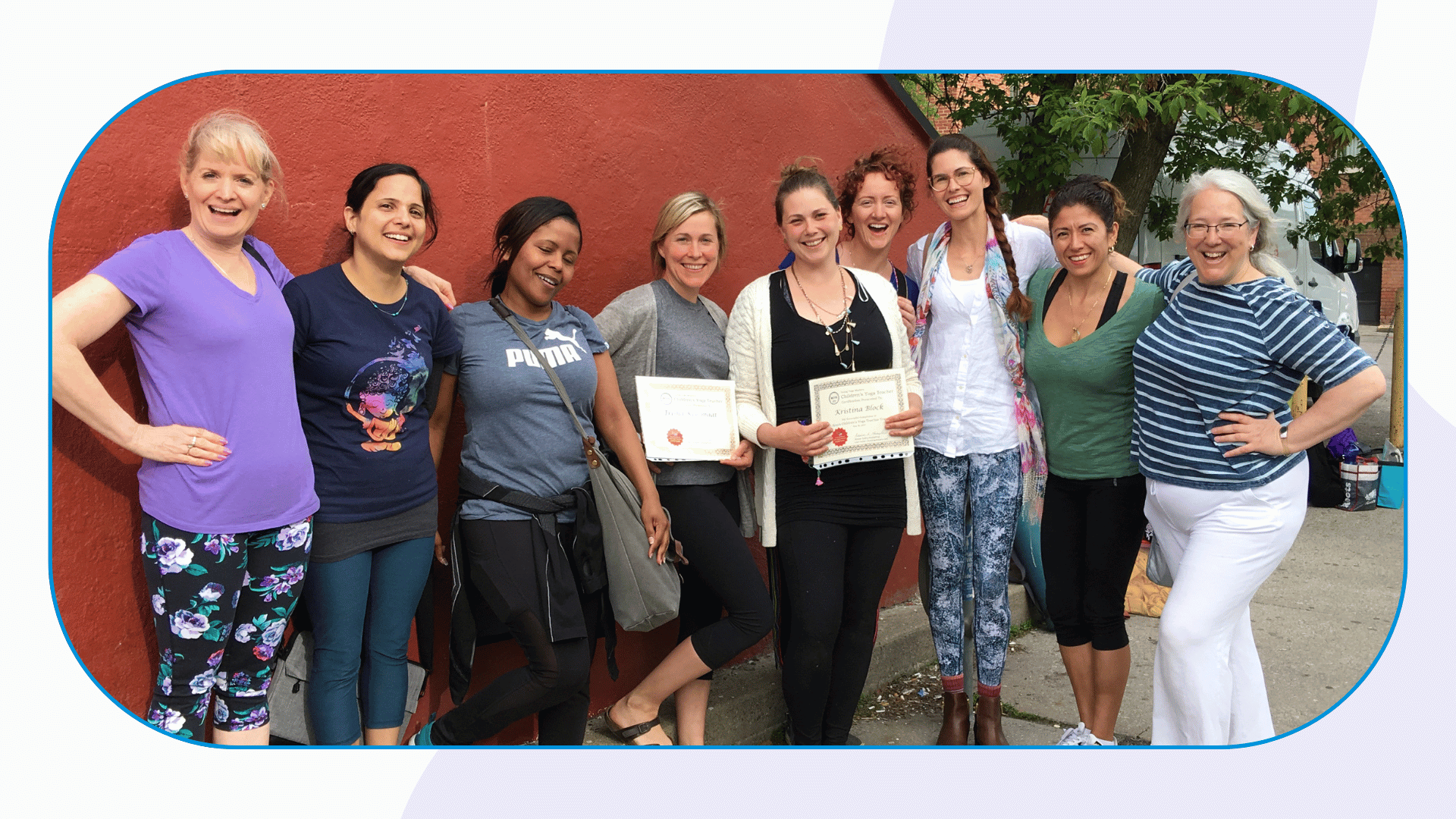 A group of 9 people stand smiling in front of a umber wall with 2 people holding graduation certificates for the kids yoga teacher training