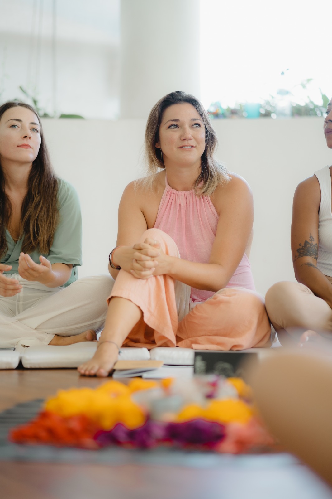 A picture of Katie from KatieHart.JustBreathe. She is facilitating a breathwork and reiki session. She is wearing a pink jumpsuit. She is sat in front of a beautiful flower alter. Katie is smiling at the other clients in the class. They are all there to welcome in more self love and mindfulness.