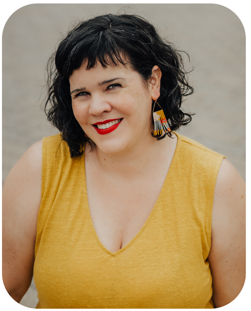 Portrait of Letitia Henville, a white lady with black-and-grey hair. Letitia is wearing beaded earrings, bright red lipstick, and a yellow sleeveless top. She is smiling broadly. 