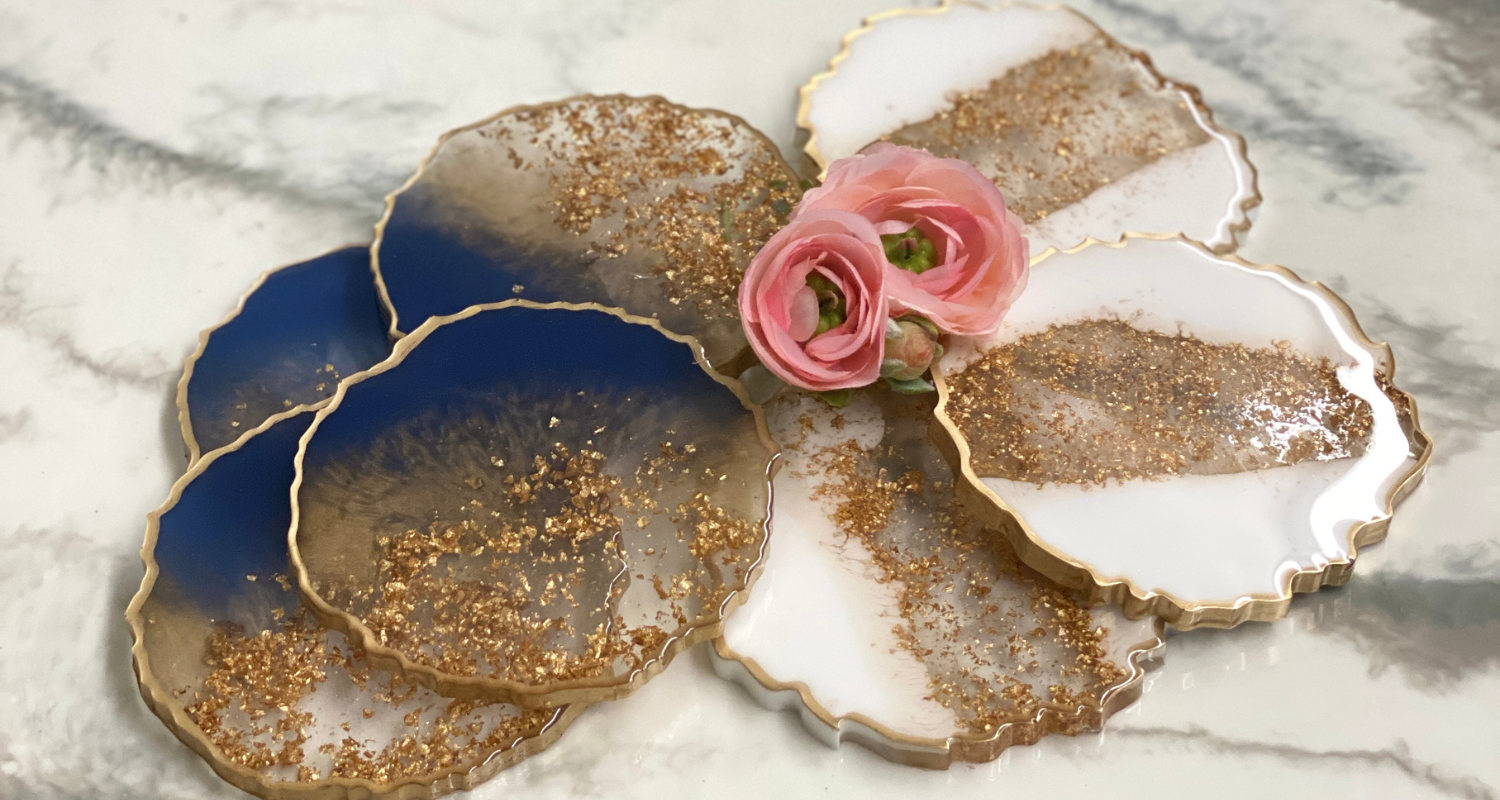 A luxurious set of resin art coasters, each boasting a unique blend of deep navy and translucent white with glittering gold leaf accents along their scalloped edges. These handcrafted pieces rest elegantly on a marble surface, adorned with a trio of delicate pink roses, adding a touch of romantic flair to the scene. The coasters exemplify the fusion of functional art with upscale decor, perfect for enhancing any table setting.