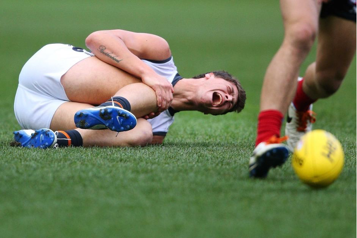 Young AFL athlete injured with an ACL tear in the AFL field while others play around him.