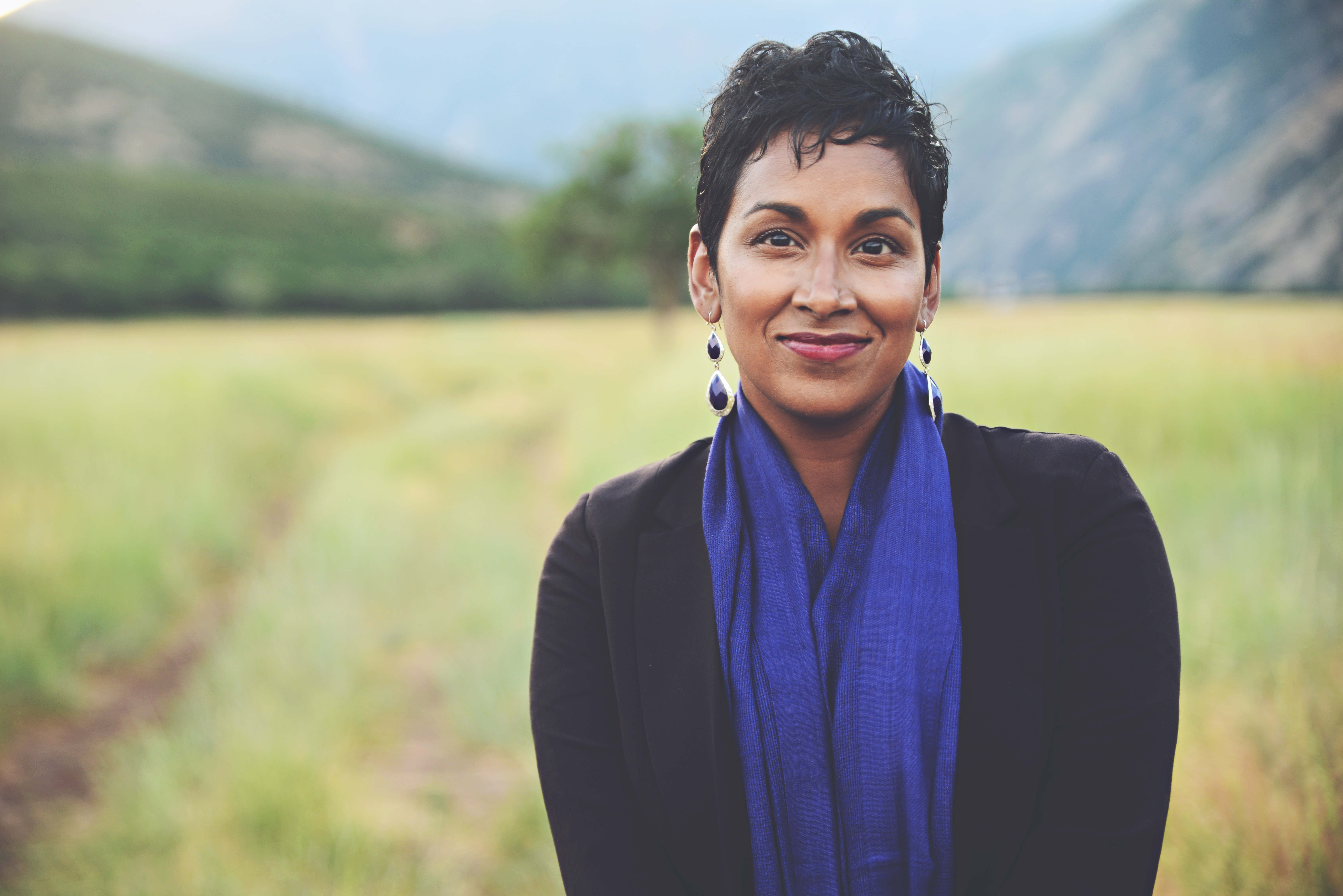 photo of Leatha, a woman of South Asian descent, with brown skin and short black wavy hair, smiling and looking directly at the camera. She is wearing a black blazer and a blue scarf.