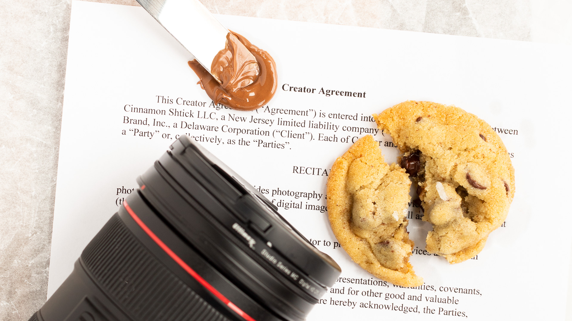 first page of a contract with a camera lease, a chocolate chip cookie broken in half, and a metal spatula with melted chocolate on top of it