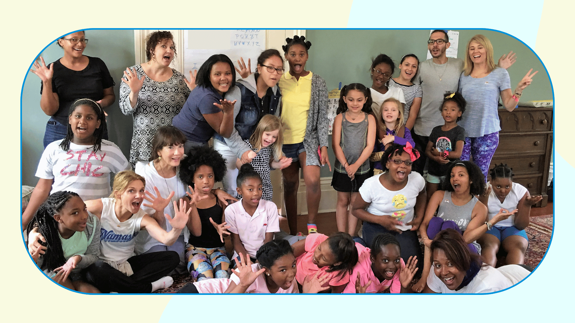 A large group of children and teachers pose making funny faces.  Some are standing and some are sitting and lying on the floor.  there is a yoga alphabet game in the background on the wall.