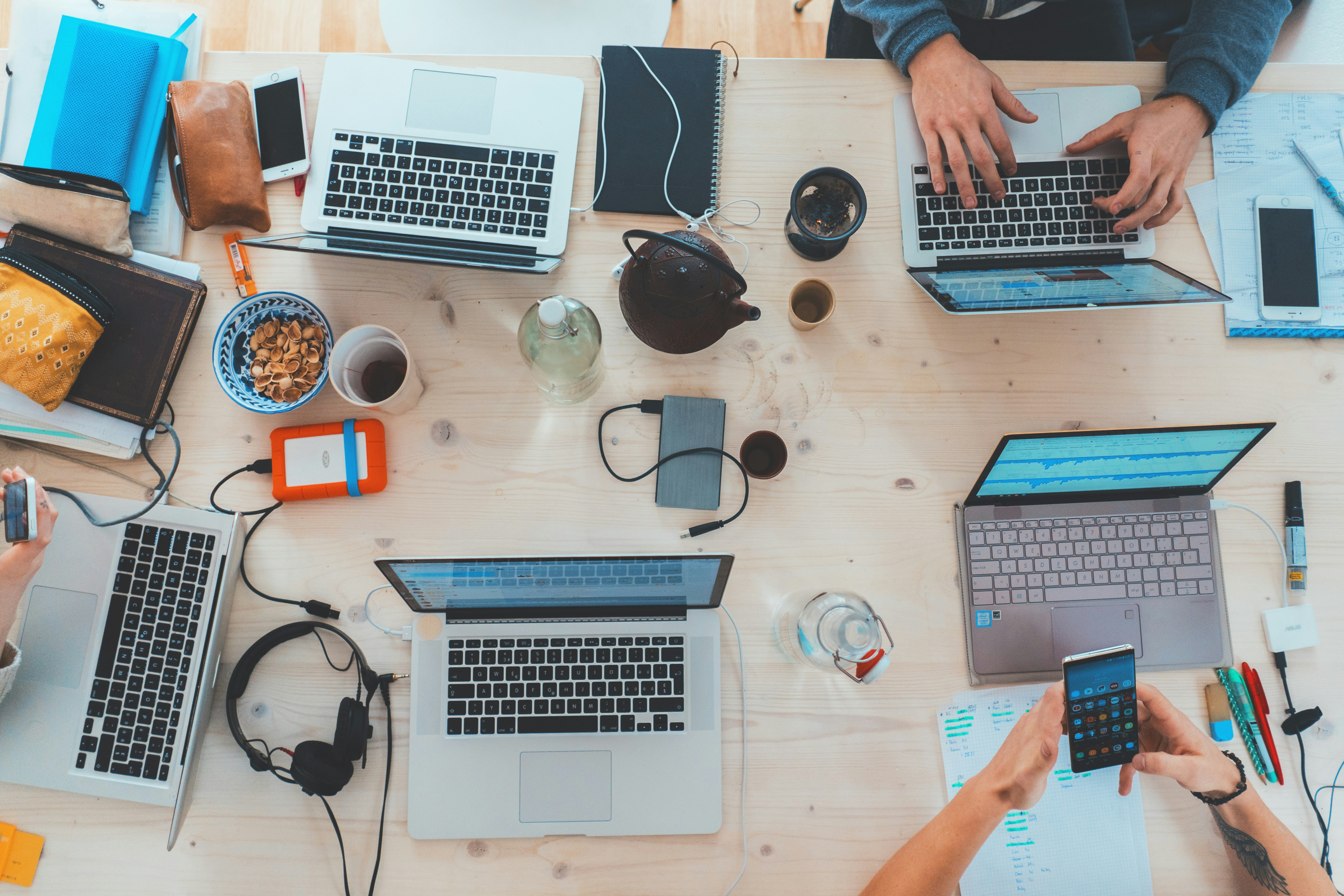 study desk with multiple laptops, charger cords, snacks and people typing