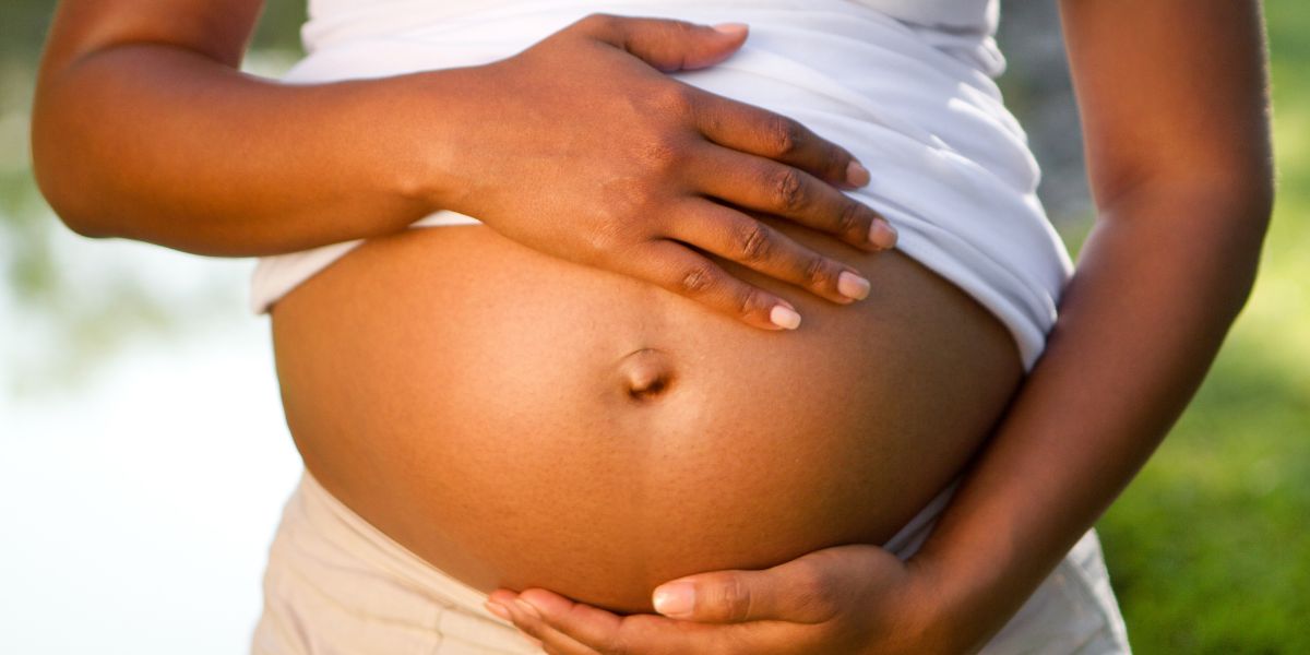 Pregnant person with dark skin and white shirt lifted to expose belly is holding belly on the top and bottom.