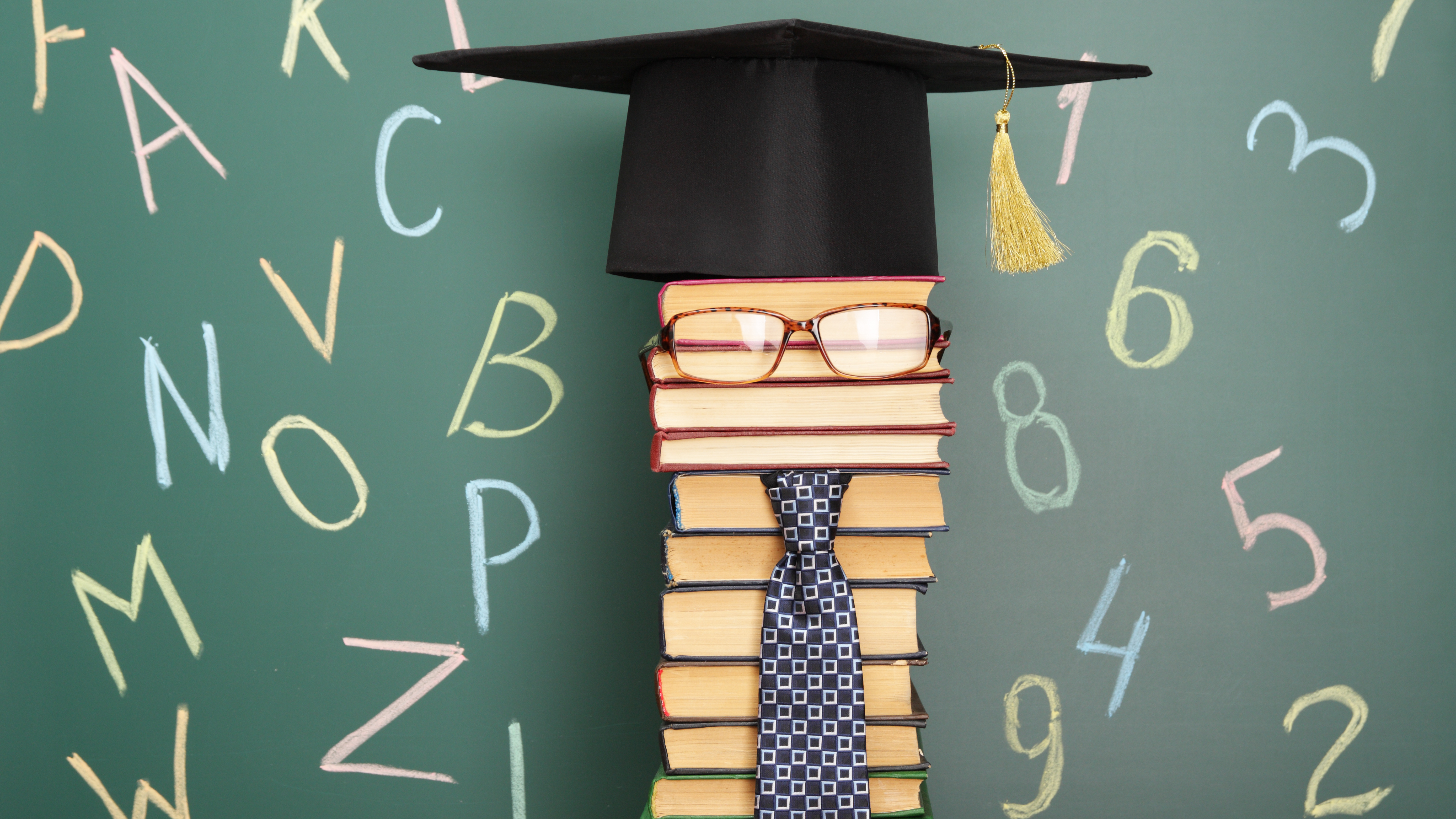 stack of books turned into a graduate cap and tie in front of chalkboard