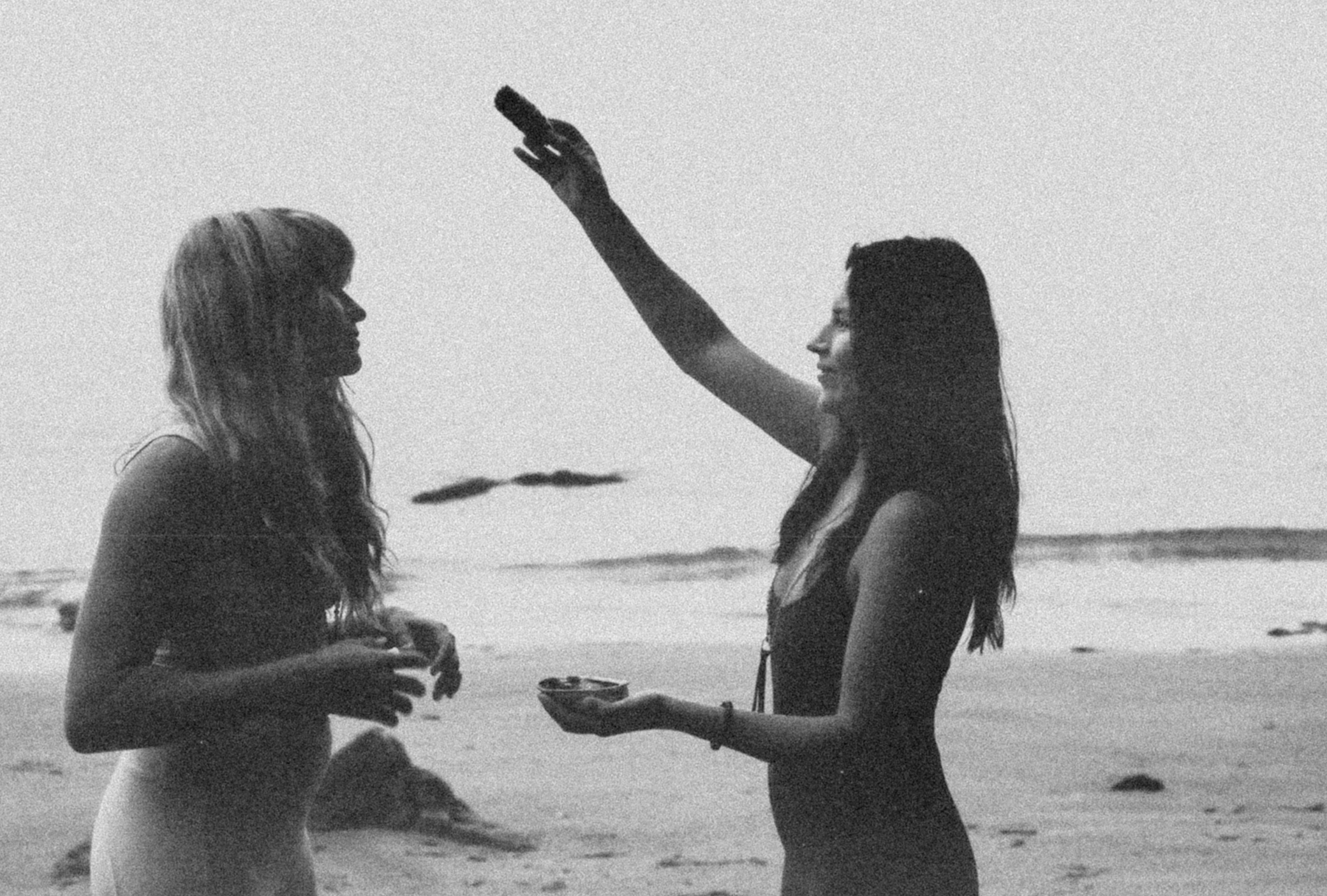 A women cleanses another women with a smoking bundle of sage on the beach. 